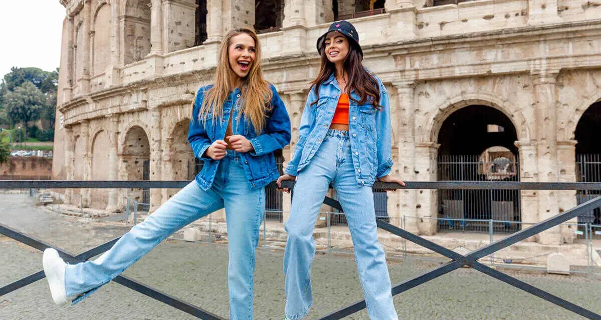 Dwie kobiety w jeans total look pozują przed Koloseum. Jedna ma rozpuszczone włosy i szeroki uśmiech, druga w kapeluszu bucket hat.